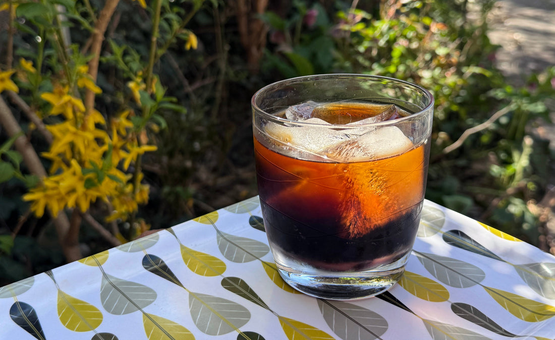 Glass of cold brew coffee over ice on a decorative gold, grey, and black tray perched in a sunny garden.