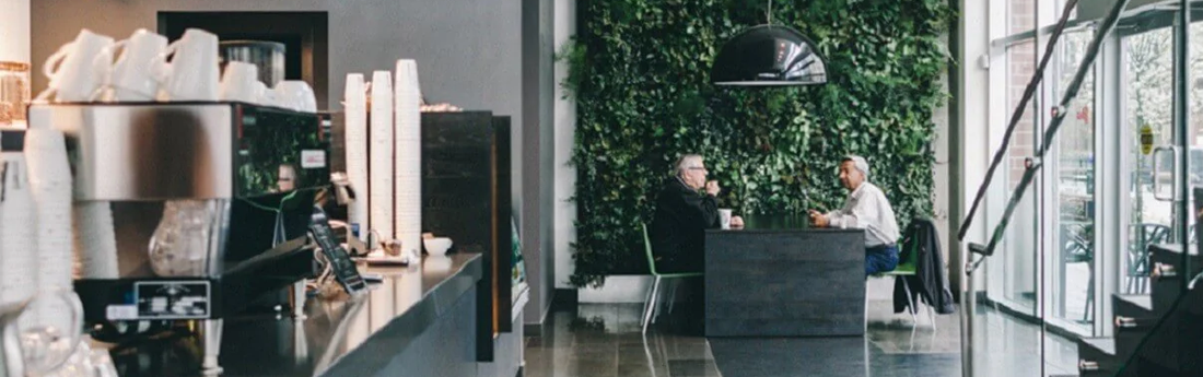 Silver espresso machine with cups on top in foreground with two people in background enjoying a meal in a grey and green restaurant space. Swiss Water Process don't let a great meal be spoiled by bad coffee