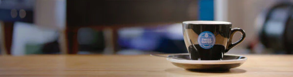 Black coffee cup with blue and white Swiss Water logo with blurred background Swiss Water Decaf Knowledge is Choice