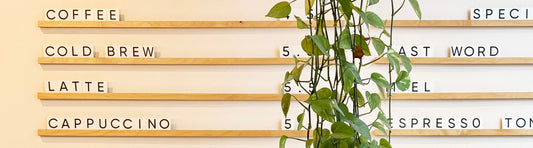 Cafe menu with various drinks in block letters on wooden rails. A green hanging plant dangles in front. Swiss Water Decaf on the Menu Welcoming Decaf Drinkers to Your Cafe