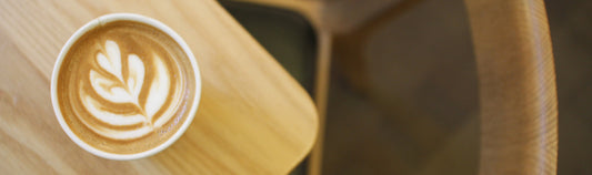 Latte with rosetta pattern on top of brown wooden table with wooden chair adjacent. Swiss Water Process Knowledge is Wellness