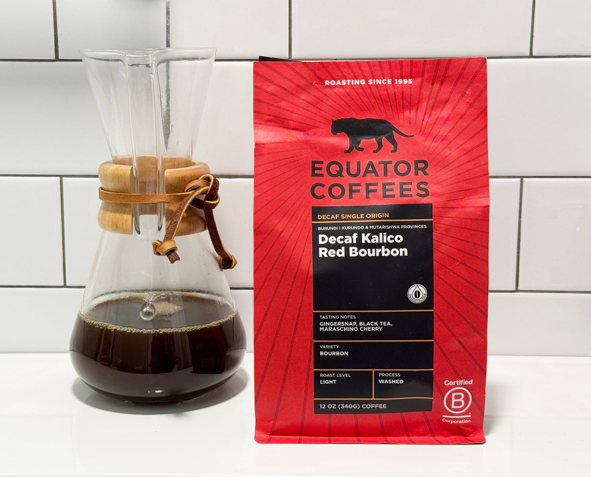 A red bag of Equator Coffees Swiss Water Decaf Kalico Red Bourbon coffee stands next to a glass pour-over coffee maker filled with dark coffee on a white tiled countertop.